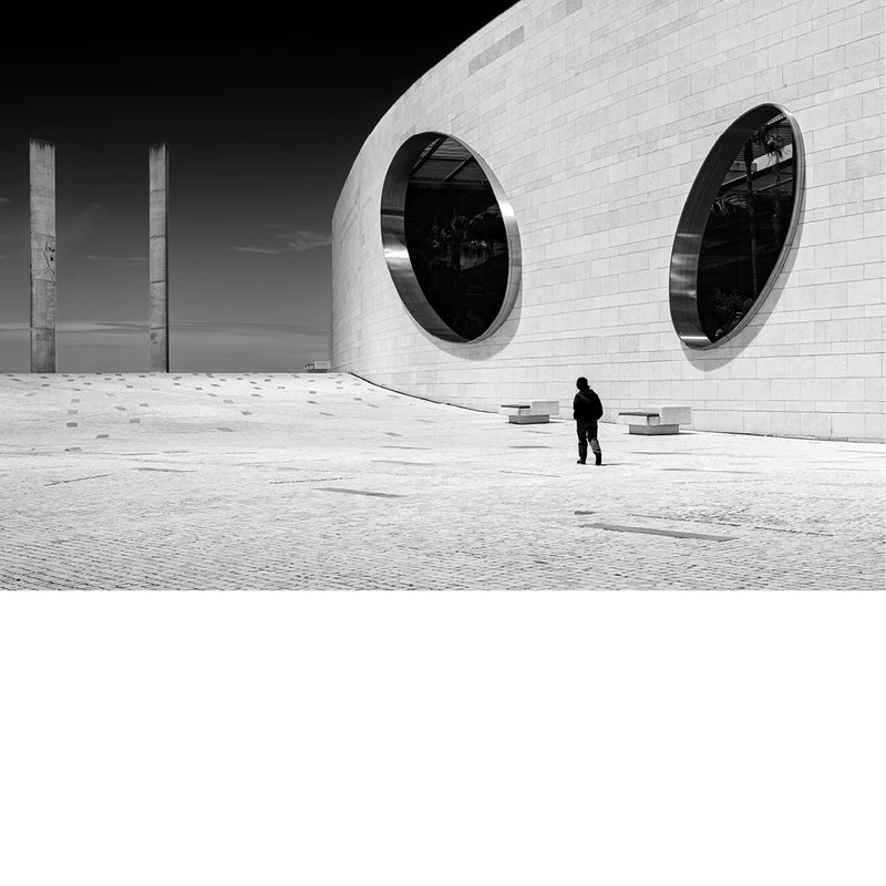 Wit gebogen gebouw van de Champalimaud Foundation in Lissabon met twee grote ronde vensters en een eenzame wandelaar op een zonovergoten plein, tegen een diepzwarte lucht.