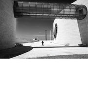 Glazen buisvormige loopbrug tussen de witte gebogen gevels van de Champalimaud Foundation in Lissabon, met een voorbijganger in het felle zonlicht en diepe slagschaduwen.