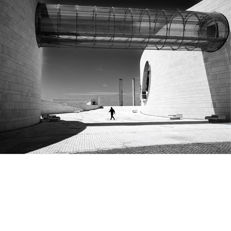 Glazen buisvormige loopbrug tussen de witte gebogen gevels van de Champalimaud Foundation in Lissabon, met een voorbijganger in het felle zonlicht en diepe slagschaduwen.