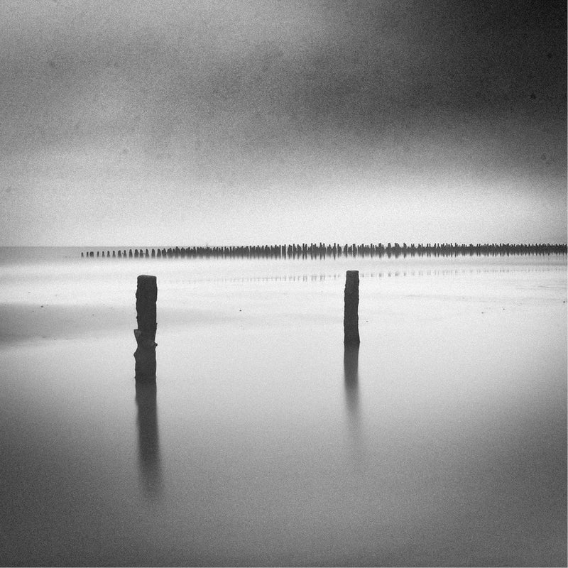Twee eenzame houten palen staan in spiegelglad water voor de kust van Wissant, met een lange rij palen in de nevel aan de horizon.