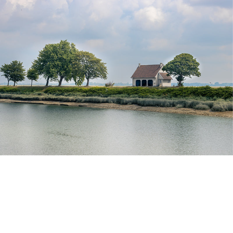 Wit huisje met rood dak tussen weelderige bomen op een groene oever in Honfleur, weerspiegeld in kalm water onder een bewolkte lucht.