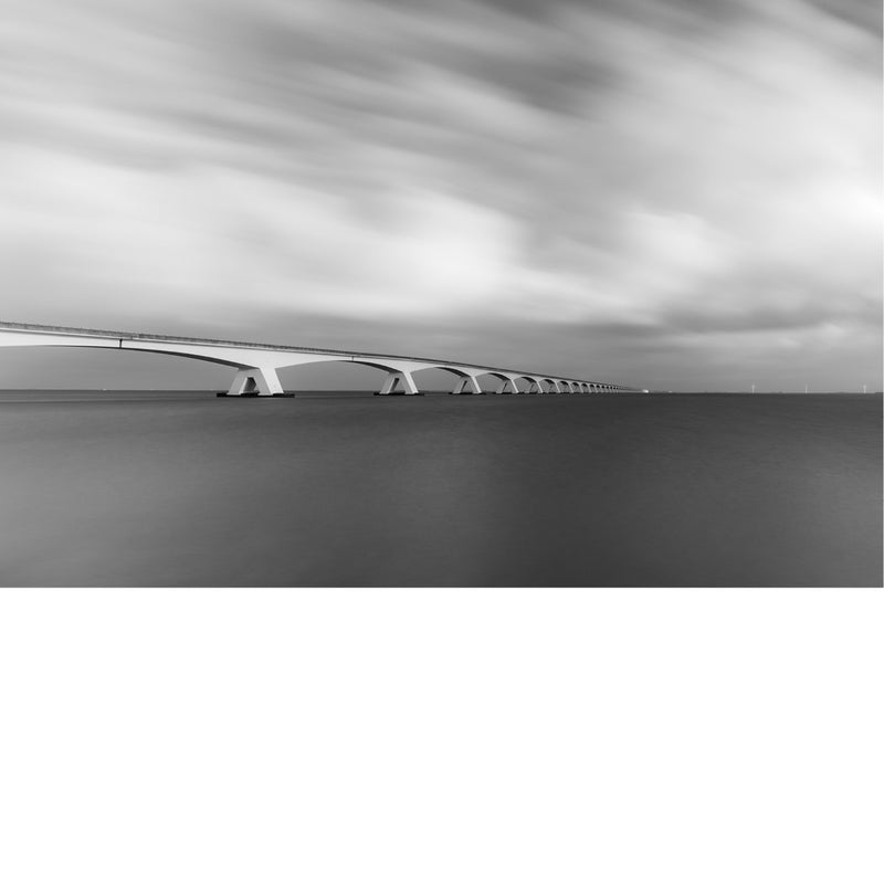 Zeelandbrug verdwijnt met haar talrijke bogen naar de horizon boven donker, glad water, onder een lucht vol voortjagende wolken.