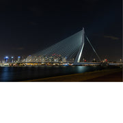 Verlichte Erasmusbrug met haar schuine pyloon en tuikabels tegen een donkere nachthemel, met de skyline van Rotterdam die in het water glinstert.