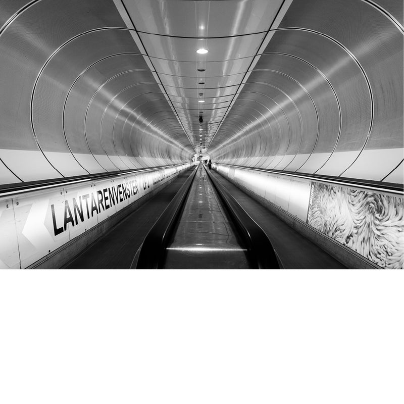 Lange, ronde gang van metrostation Wilhelminaplein in Rotterdam, met rolband en lichtlijnen die in perspectief naar een klein lichtpunt in de verte lopen.