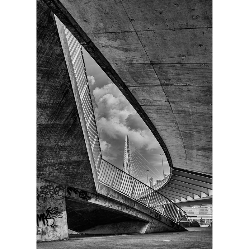 Driehoekige opening in een betonnen constructie kadert de witte pyloon van de Erasmusbrug in Rotterdam en stapelwolken, met graffiti op de voorgrond.