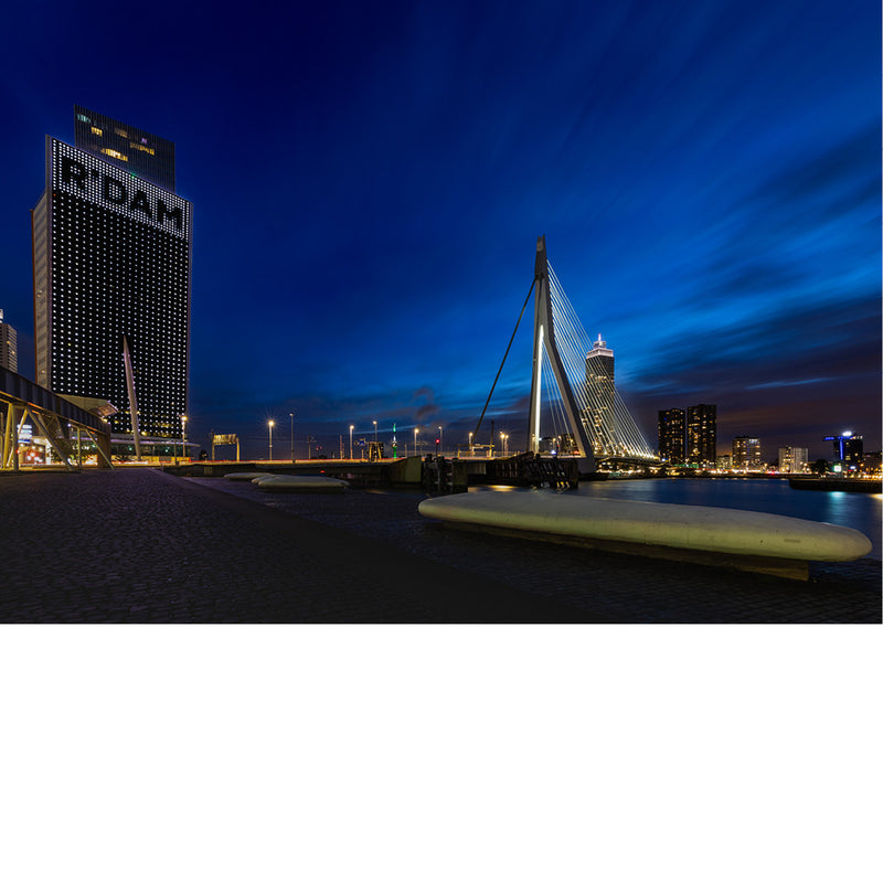 Erasmusbrug en verlichte hoogbouw met het opschrift R'DAM in het diepblauwe avondlicht, met strepen van bewegende wolken.