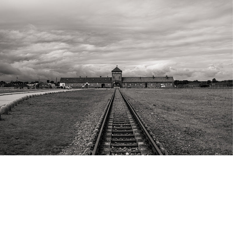 Spoorlijn loopt recht naar het poortgebouw van Auschwitz, met een wijd grasveld onder een grijze wolkenlucht.