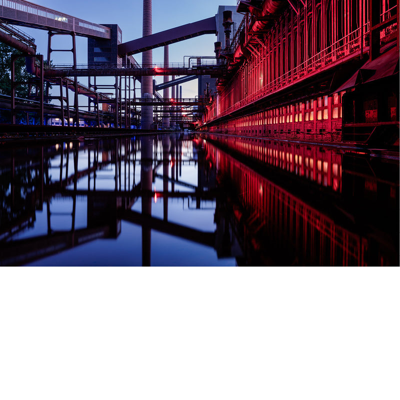 Cokesfabriek van Zollverein in Essen met rood verlichte gevel en stalen transportbanden, weerspiegeld in een waterbekken tijdens het blauwe uur.