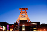 Oranje verlichte schachtbok van Zollverein in Essen boven bakstenen gebouwen, tegen een diepblauwe avondlucht.