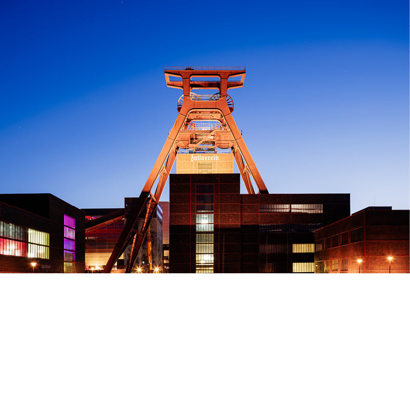 Oranje verlichte schachtbok van Zollverein in Essen boven bakstenen gebouwen, tegen een diepblauwe avondlucht.