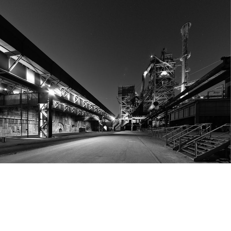 Verlichte hoogoven en een lange overdekte hal in het Landschaftspark Duisburg-Nord langs een verlaten weg onder een donkere nachthemel.