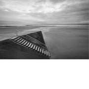 Een minimalistisch zwart-wit beeld van een betonnen trap met metalen leuning die uitkomt op een uitgestrekt, leeg strand bij een bewolkte hemel.