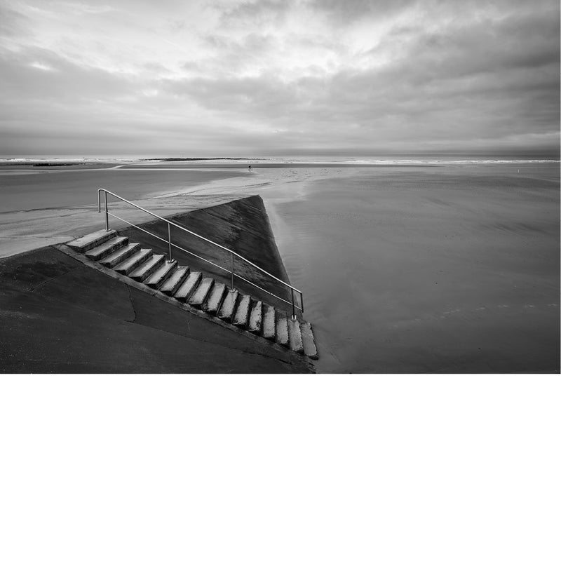 Een minimalistisch zwart-wit beeld van een betonnen trap met metalen leuning die uitkomt op een uitgestrekt, leeg strand bij een bewolkte hemel.