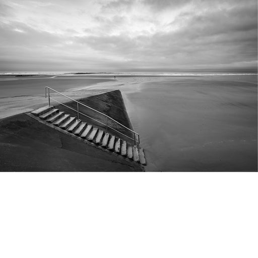 Een minimalistisch zwart-wit beeld van een betonnen trap met metalen leuning die uitkomt op een uitgestrekt, leeg strand bij een bewolkte hemel.