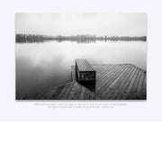Een melancholisch landschap in zwart-wit. Een houten steiger strekt zich uit in kalm, spiegelglad water, omhuld door lichte mist op de achtergrond.