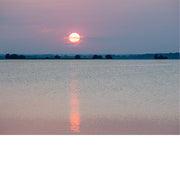 Gloeiende zon hangt laag boven een rustig wateroppervlak en werpt een lange rode weerspiegeling over het water, met een donkere bomenrand aan de horizon.