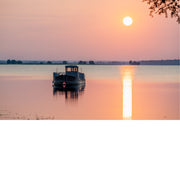 Aangemeerde boot ligt op het water terwijl een gloeiende zon het tafereel in oranje en roze licht zet, met een lange lichtreflectie op het wateroppervlak.