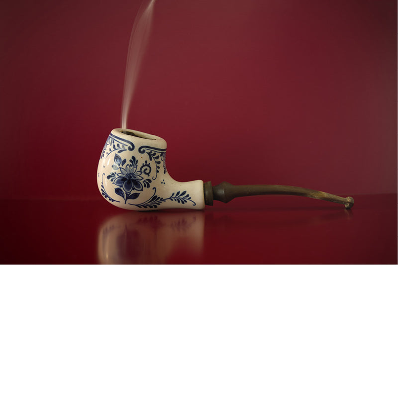 Een pijp in Delfts blauw waaruit een bescheiden rookpluim komt; tegen een rode achtergrond