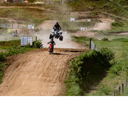 Quad die hoog boven een stoffige crossbaan springt, terwijl een jonge rijder op een rode crossmotor de heuvel op komt.