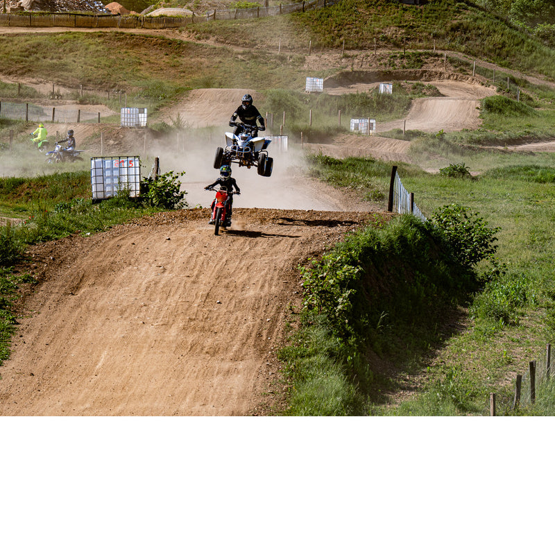 Quad die hoog boven een stoffige crossbaan springt, terwijl een jonge rijder op een rode crossmotor de heuvel op komt.