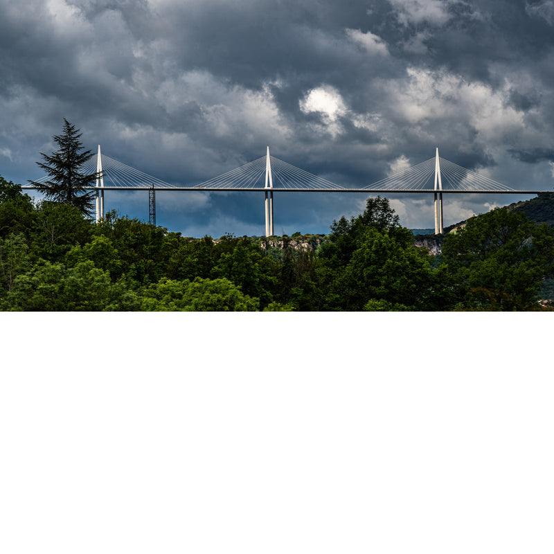 Witte pylonen van een lange tuibrug steken boven een groene heuvelrug uit, onder een zwaar bewolkte lucht met een enkele lichtplek.