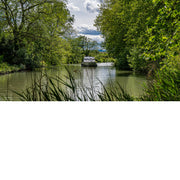 Plezierboot vaart over een kanaal tussen dichtbegroeide oevers, met rietstengels op de voorgrond en wolken boven de bomen.