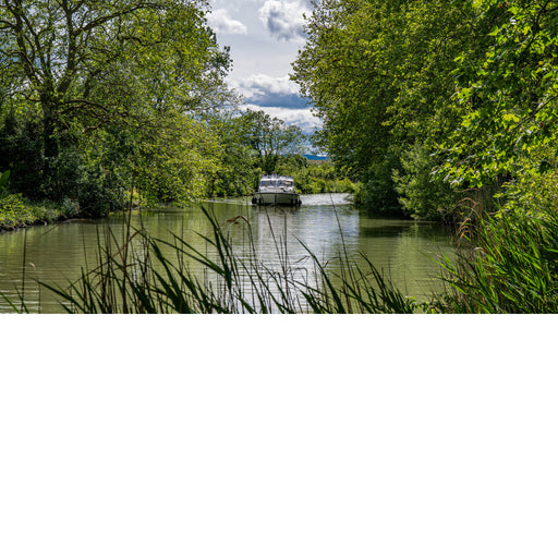 Plezierboot vaart over een kanaal tussen dichtbegroeide oevers, met rietstengels op de voorgrond en wolken boven de bomen.