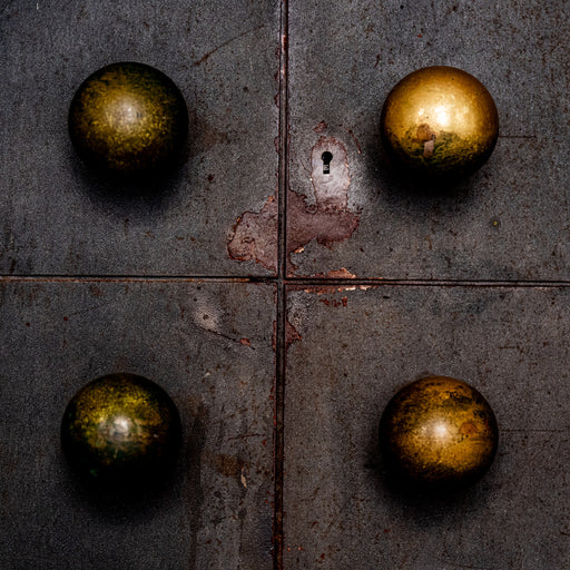 Vier koperen bolknoppen en een sleutelgat op een donkere, verweerde metalen deur met roestplekken.