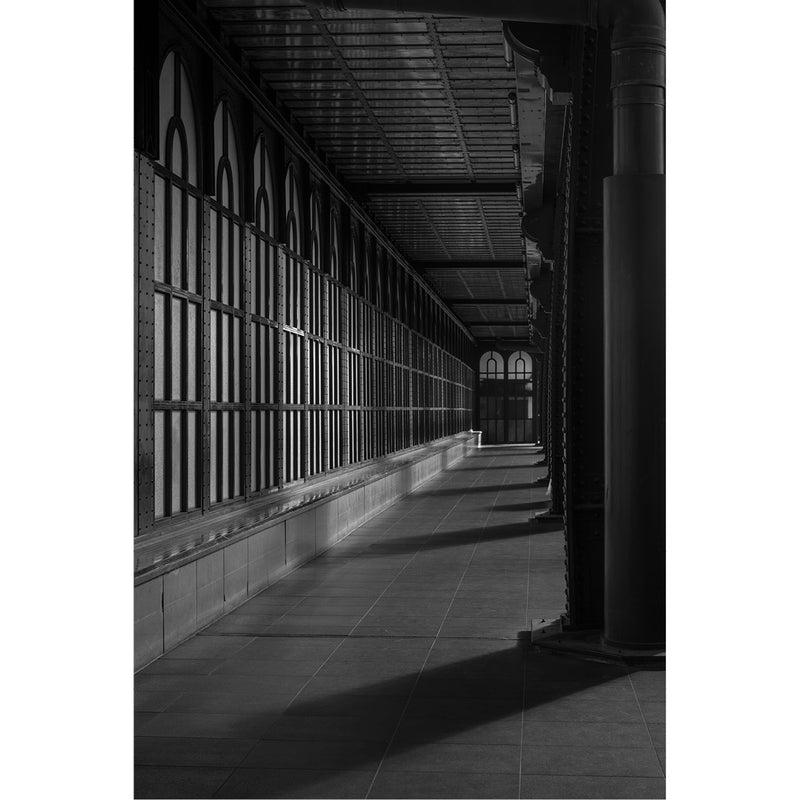 Lange industriële galerij met repeterende stalen vensters, waar strepen van licht en schaduw over de vloer vallen.