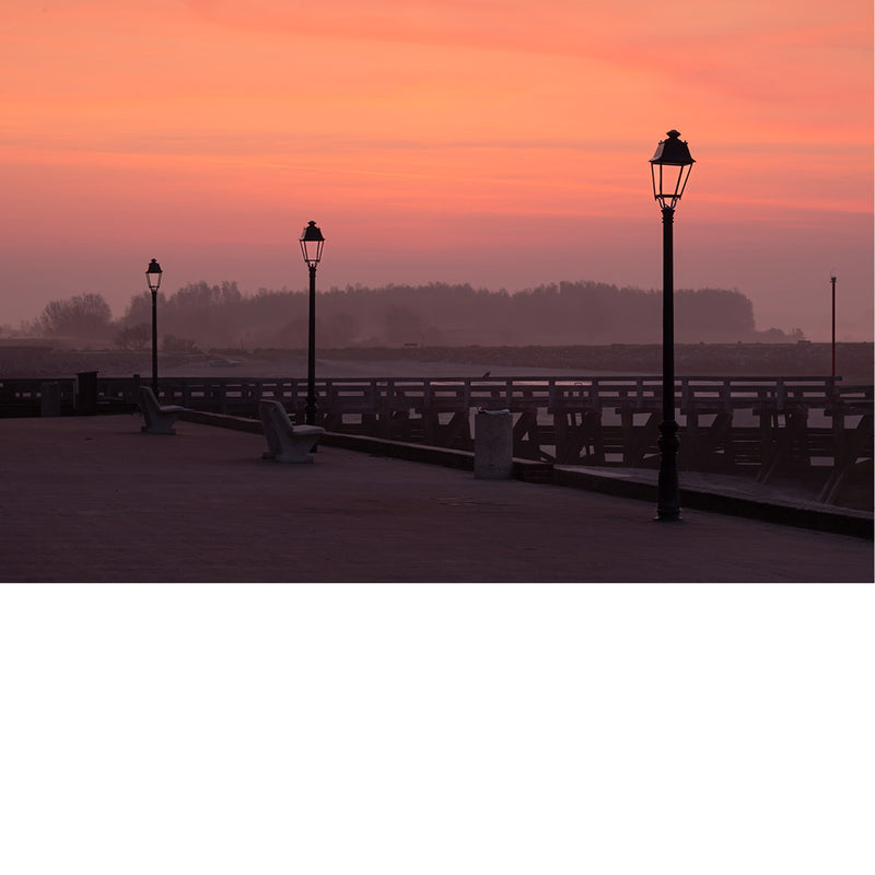 Straatlantaarns en bankjes staan op een lege promenade aan de Baai van de Somme, tegen een roze-oranje avondlucht met opkomende mist.