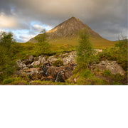 Kleine watervalletjes stromen tussen rotsblokken op de voorgrond, met de karakteristieke piek van Buachaille Etive Mor op de achtergrond onder een bewolkte lucht.
