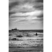Een eenzame zwemmer trotseert de woelige Noordzee terwijl een patrouilleboot onder een dramatisch wolkendek de horizon doorkruist. Deze sfeervolle kustfoto, vastgelegd in zwart-wit, roept gevoelens op van eenzaamheid, vrijheid en de tijdloze kracht van de zee.