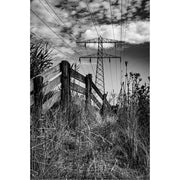 Een verweerd houten hek en wild grasland vormen de voorgrond van dit contrastrijke landschap. Daarboven rijst een imposante hoogspanningsmast op tegen een dramatische wolkenlucht. De grillige natuur en strakke lijnen van de elektriciteitsdraden creëren een intrigerend samenspel tussen rust, ruigheid en menselijke infrastructuur.