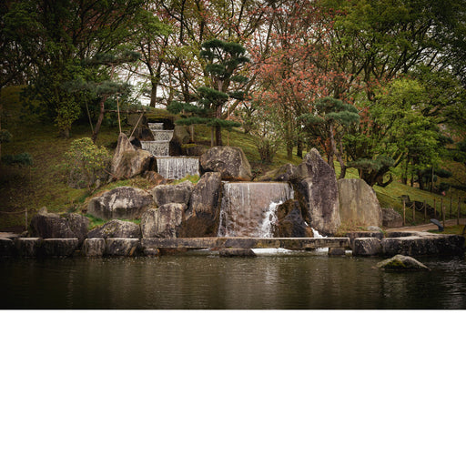 Trapsgewijze waterval tussen grote rotsblokken en gesnoeide dennen, die uitkomt in een stille vijver in de Japanse tuin van Hasselt.