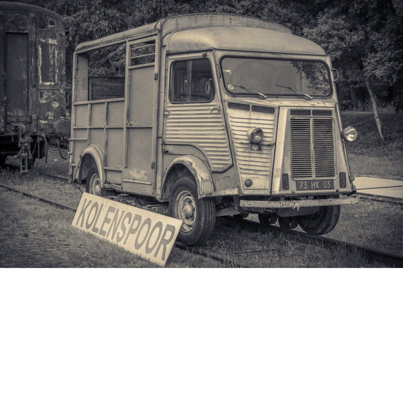 Verweerde oude Citroën-bestelwagen op de sporen bij het oude stationnetje van As, met een bord Kolenspoor en een roestige treinwagon ernaast.