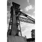Geklonken stalen schachtbok van C-mine in Genk van onderen gezien, met vakwerkliggers tegen een lucht met stapelwolken.