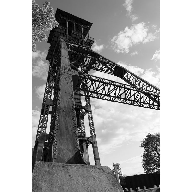 Geklonken stalen schachtbok van C-mine in Genk van onderen gezien, met vakwerkliggers tegen een lucht met stapelwolken.