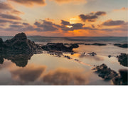 Een zonsondergang aan de kust, met de zon die ondergaat aan de horizon. De lucht is gevuld met oranje, roze en paarse tinten, die worden weerspiegeld in het water op de voorgrond. Rotsformaties zijn zichtbaar langs de kustlijn, met de golven die zachtjes over de rotsen slaan.