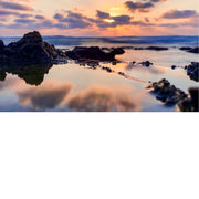 Gestileerd beeld van een zonsondergang aan de kust, met de zon die ondergaat aan de horizon. De lucht is gevuld met oranje, roze en paarse tinten, die worden weerspiegeld in het water op de voorgrond. Rotsformaties zijn zichtbaar langs de kustlijn, met de golven die zachtjes over de rotsen slaan.