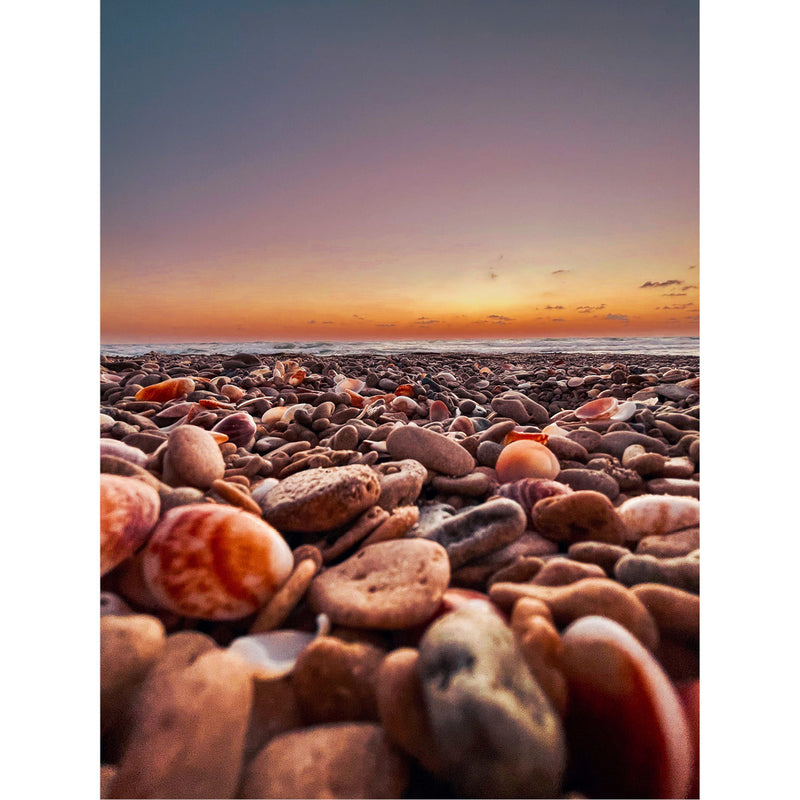 Close-up van een strand bezaaid met kiezels en schelpen bij zonsondergang. De zon is al onder, maar de hemel is nog steeds verlicht met een zachte oranje en paarse gloed.