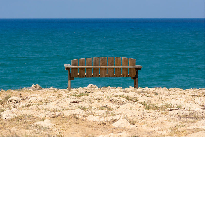 Een houten bankje op een rotsachtige kustlijn met uitzicht op een uitgestrekte, turquoise zee onder een helderblauwe lucht.