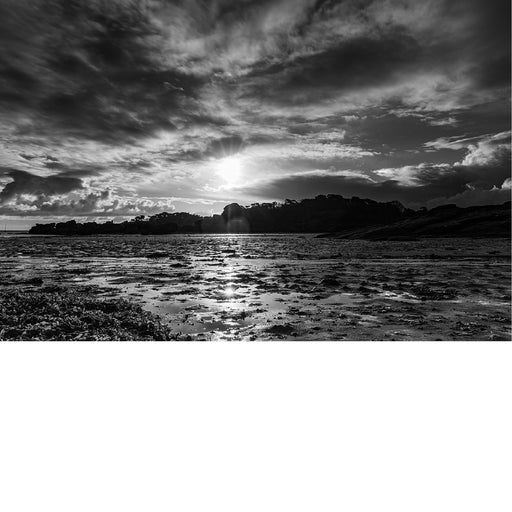 Felle zonnestralen breken door een dramatische wolkenlucht boven een wijds wad met rotsen en zeewier, in zwart-wit.