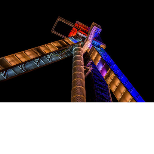 De iconische schachtbokken van C-Mine in Genk worden 's avonds sfeervol verlicht en steken krachtig af tegen de donkere hemel. Vanuit een laag standpunt benadrukt de compositie de indrukwekkende hoogte en industriële architectuur van dit voormalige mijncomplex.
