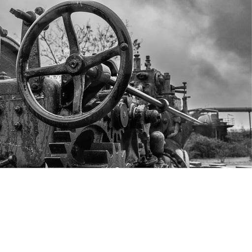 Zwart-witfoto van een oude industriële machine met tandwielen en een groot stuurwiel, buitenomgeving.
