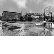 Zwart-witfoto van een verlaten industrieterrein met metalen brug en natte grond