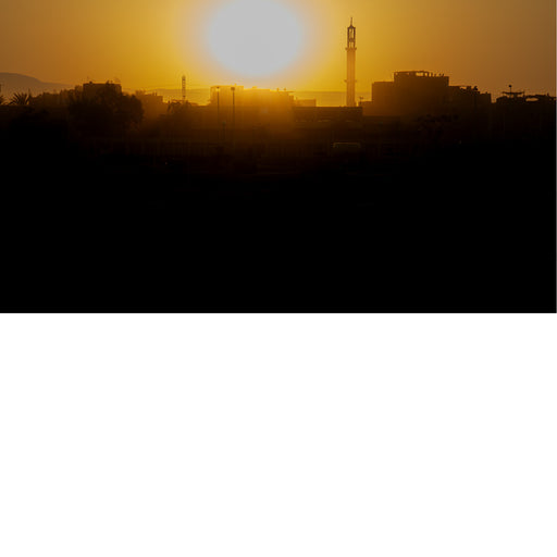 Warme oranje zonsondergang boven een stad met minaret en gebouwen in silhouet, tegen een bergachtige achtergrond.