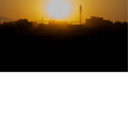 Warme oranje zonsondergang boven een stad met minaret en gebouwen in silhouet, tegen een bergachtige achtergrond.