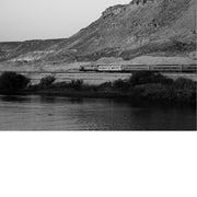 Zwart-witfoto van een lange goederentrein langs een rotsachtige heuvel, met rivier op de voorgrond.