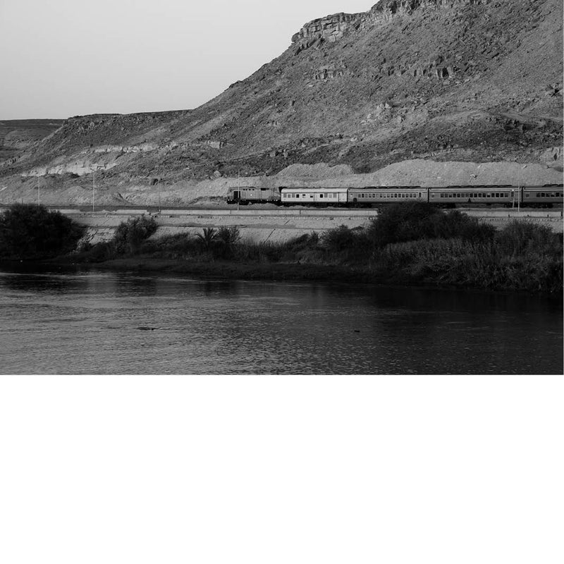 Zwart-witfoto van een lange goederentrein langs een rotsachtige heuvel, met rivier op de voorgrond.