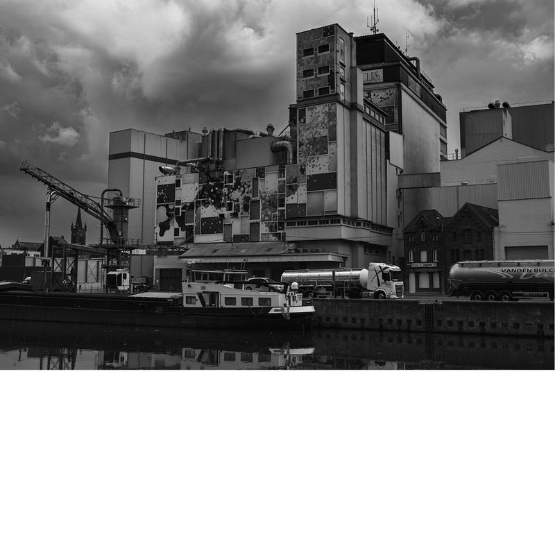 Zwart-witfoto van een grote fabriek met silo’s en pijpleidingen, vrachtwagens en boten aan een kanaal.